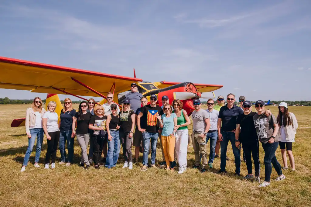 Uczestnicy pozują do zdjęcia grupowego na lotnisku, mając w tle mały samolot ustawiony przy pasie startowym.