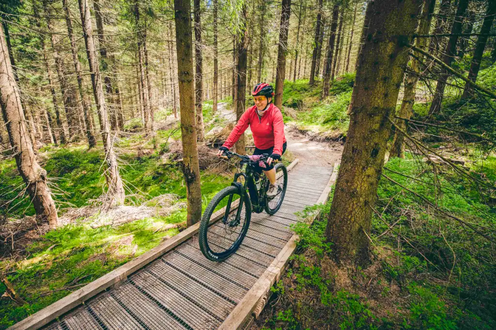 Event z serii Piąty Żywioł Ziemia, przedstawiający uczestniczkę jadącą rowerem po leśnej ścieżce podczas górskiej trasy.