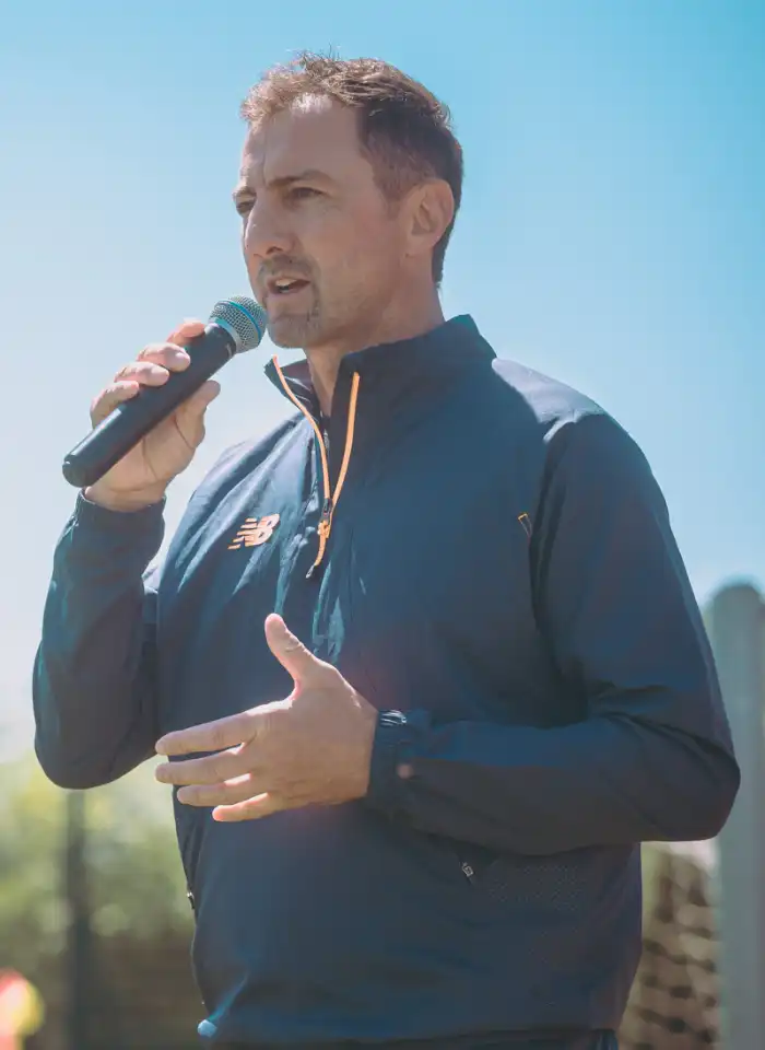 Jerzy Dudek przemawia do zawodników, trzymając mikrofon i motywując uczestników przed kolejną częścią sportowego wydarzenia.