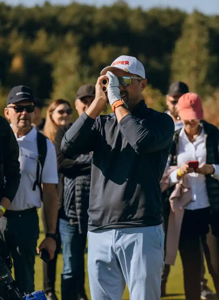 Jerzy Dudek obserwuje pole golfowe przez lunetę, analizując odległość i warunki gry podczas turnieju.