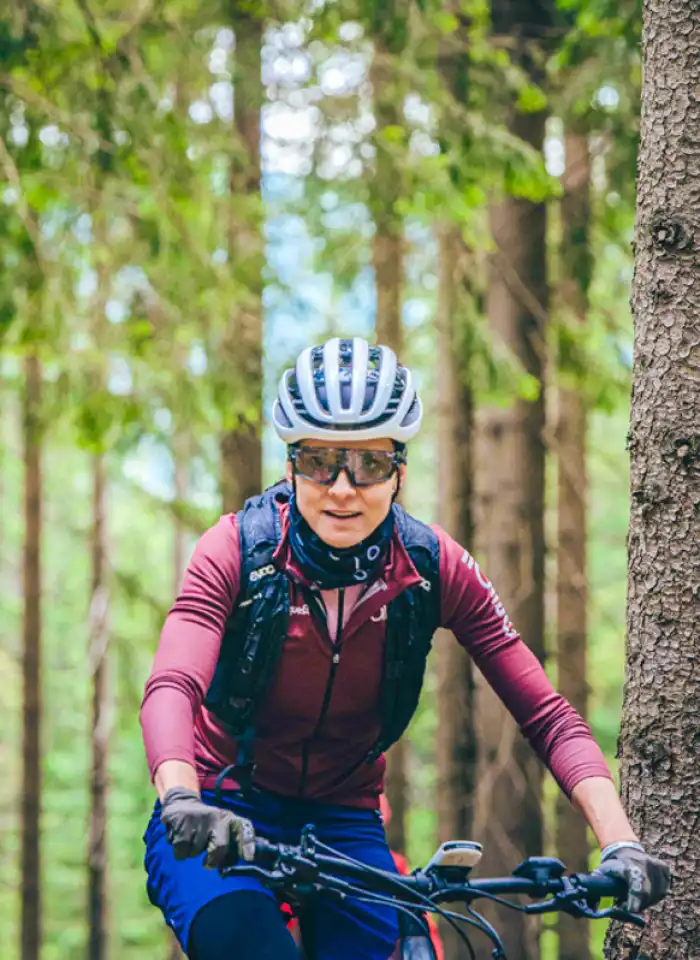 Maja Włoszczowska stoi przodem do obiektywu w kolarskim stroju, pozując na tle zielonego, leśnego otoczenia.