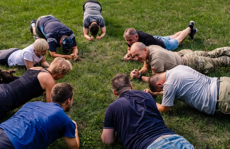 Grupa uczestników leży na trawie, rozgrzewając się do ćwiczeń z Krzysztofem Przepiórką.