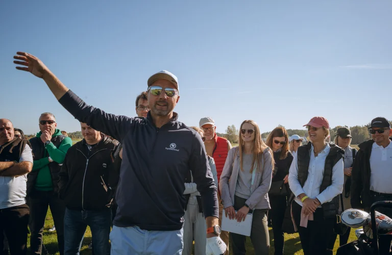 Jerzy Dudek podnosi rękę w geście powitania, motywując uczestników podczas spotkania na polu golfowym.