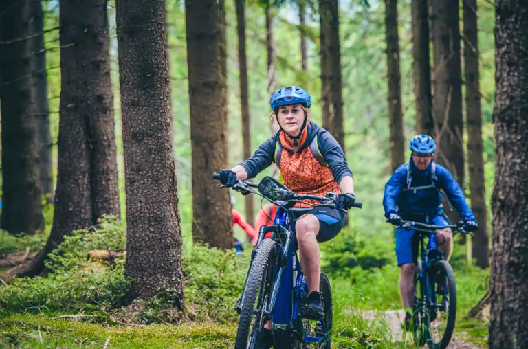 Rowerzyści jadą leśną ścieżką w wąskim pasie między drzewami, kontynuując trasę eventu terenowego.