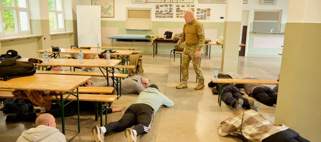 Uczestnicy szkolenia leżą na podłodze sali podczas ćwiczenia z napastnikiem, w tle instruktor