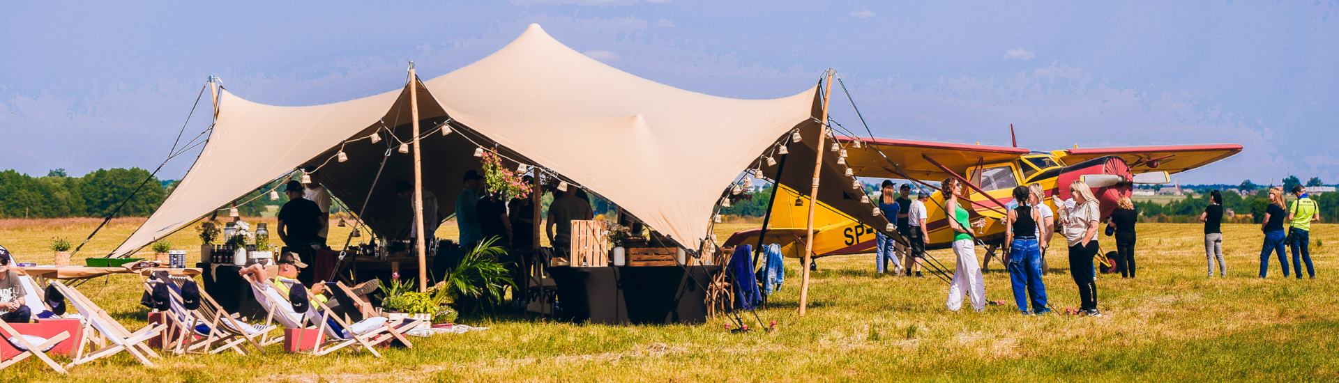Event z serii Piąty Żywioł Powietrze z Sebastianem Kawą, gdzie uczestnicy stoją pod namiotem na lotnisku obok samolotów ultralekkich.